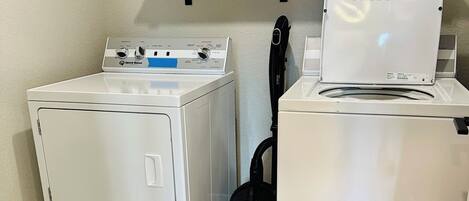 A laundry area with two white washing machines, a shelf with cleaning supplies, and a vacuum cleaner.