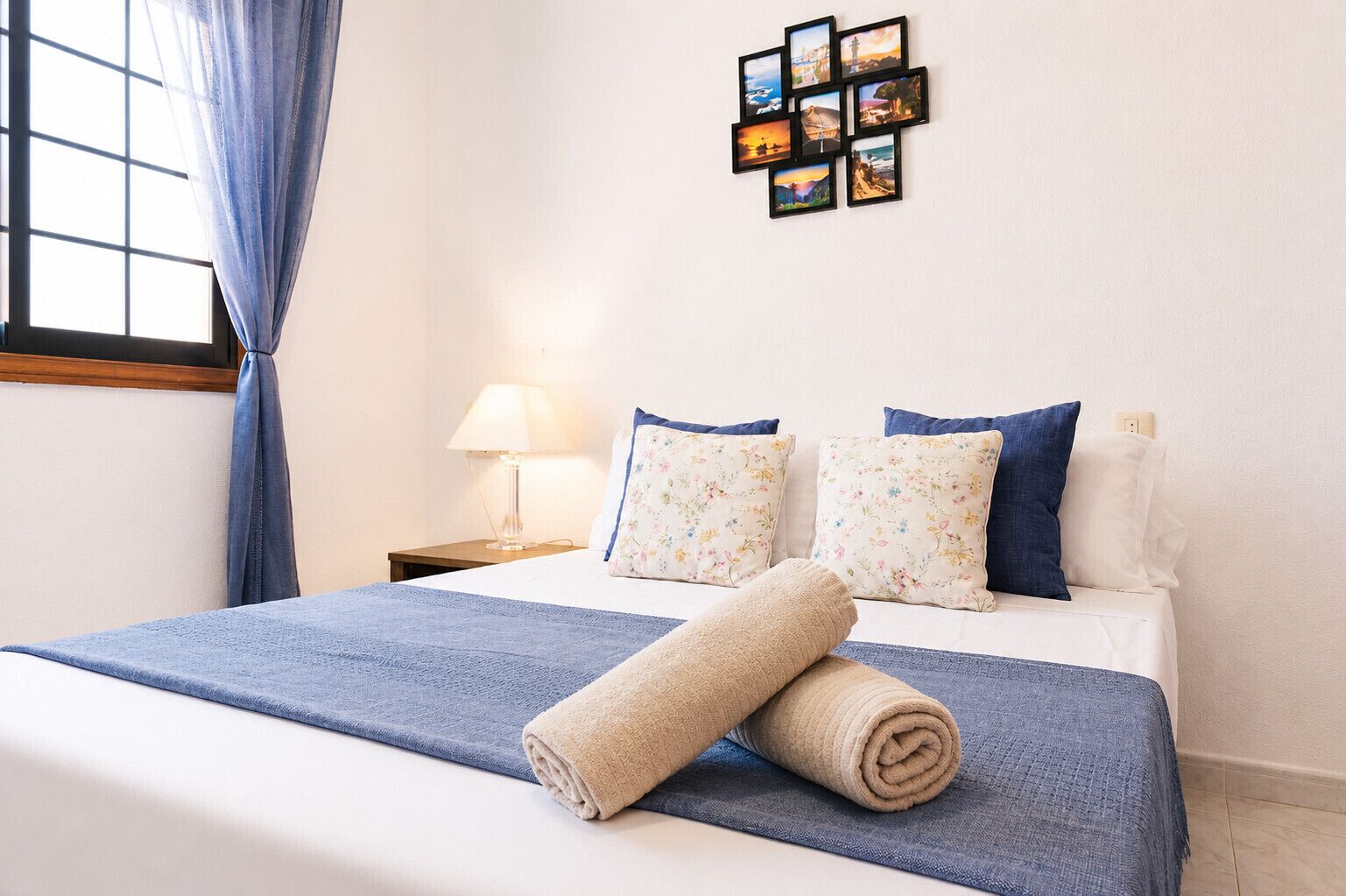 Un lit bien fait, avec une couvre-lit bleue et deux oreillers, un collage de photos accroché au mur, une lampe de chevet et une fenêtre avec des rideaux.