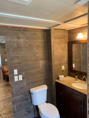 A bathroom with a wooden wall, a white toilet, a sink with a mirror, and a towel rack. - Minutes to town or very peaceful woods retreat! Close to trails and golf. (Lewiston)