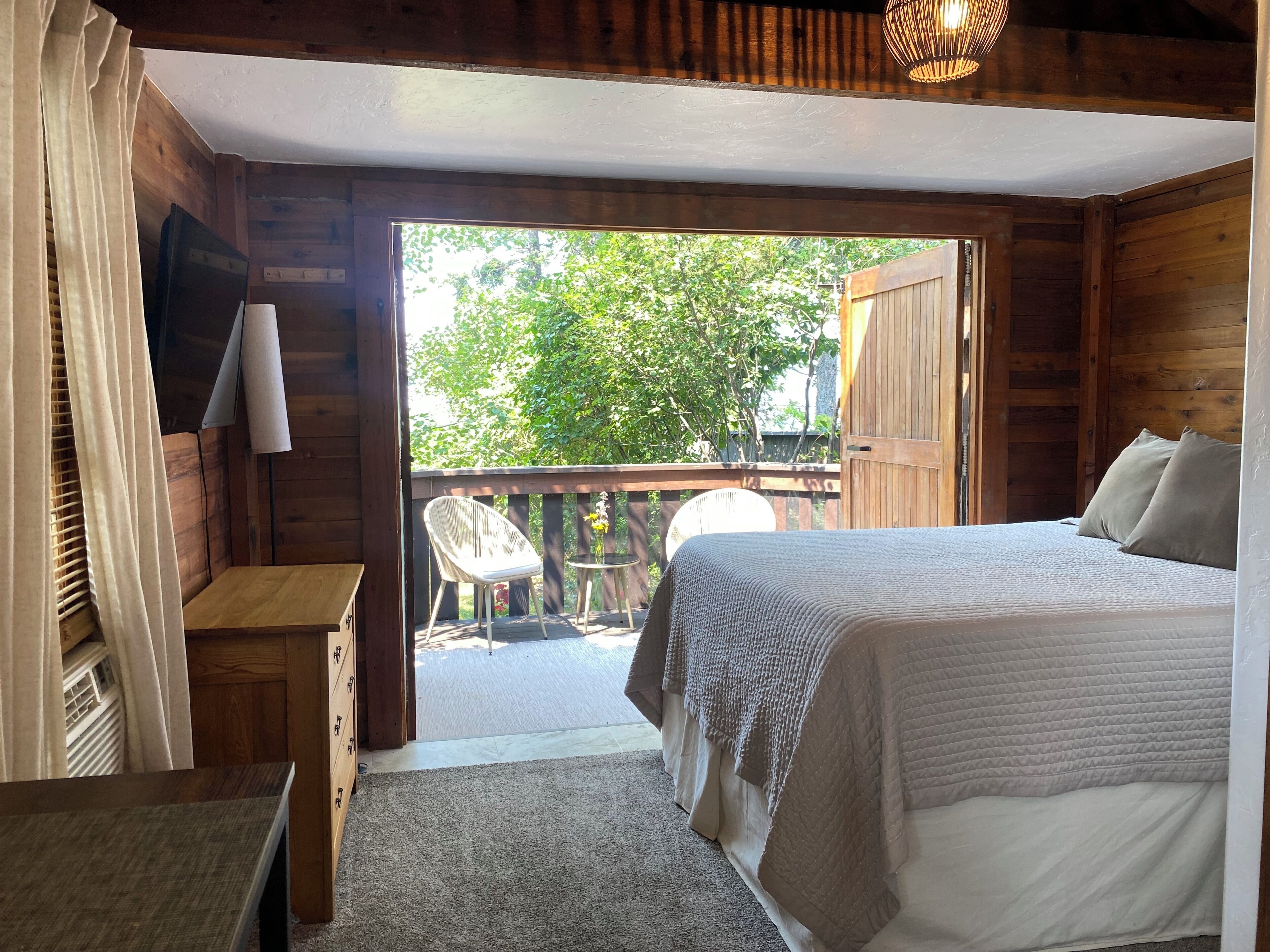 A bedroom with a bed, a desk, and a chair. There is a balcony with a view of trees.