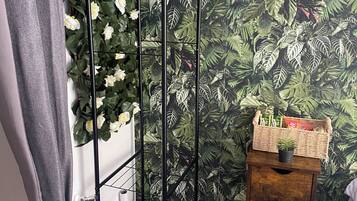 Een kamer met een zwarte metalen plantenstandaard tegen een muur met een tropisch bladpatroon, een houten bijzettafeltje met een mand, en een grijze gordijn.