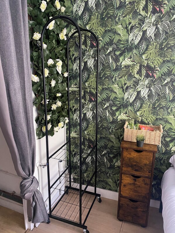 A room with a black metal plant stand against a wall with a tropical leaf pattern, a wooden side table with a basket, and a grey curtain.