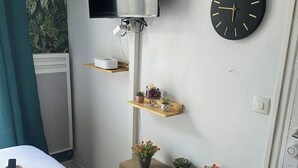 A hotel room with a bed, a mounted TV, a wall clock, and decorative shelves with plants.