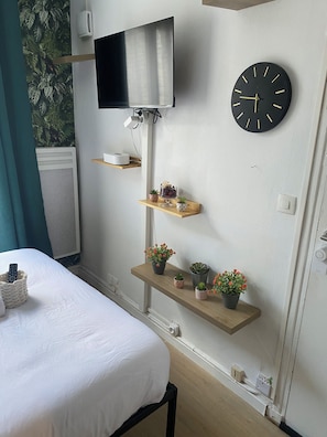 A hotel room with a bed, a mounted TV, a wall clock, and decorative shelves with plants.