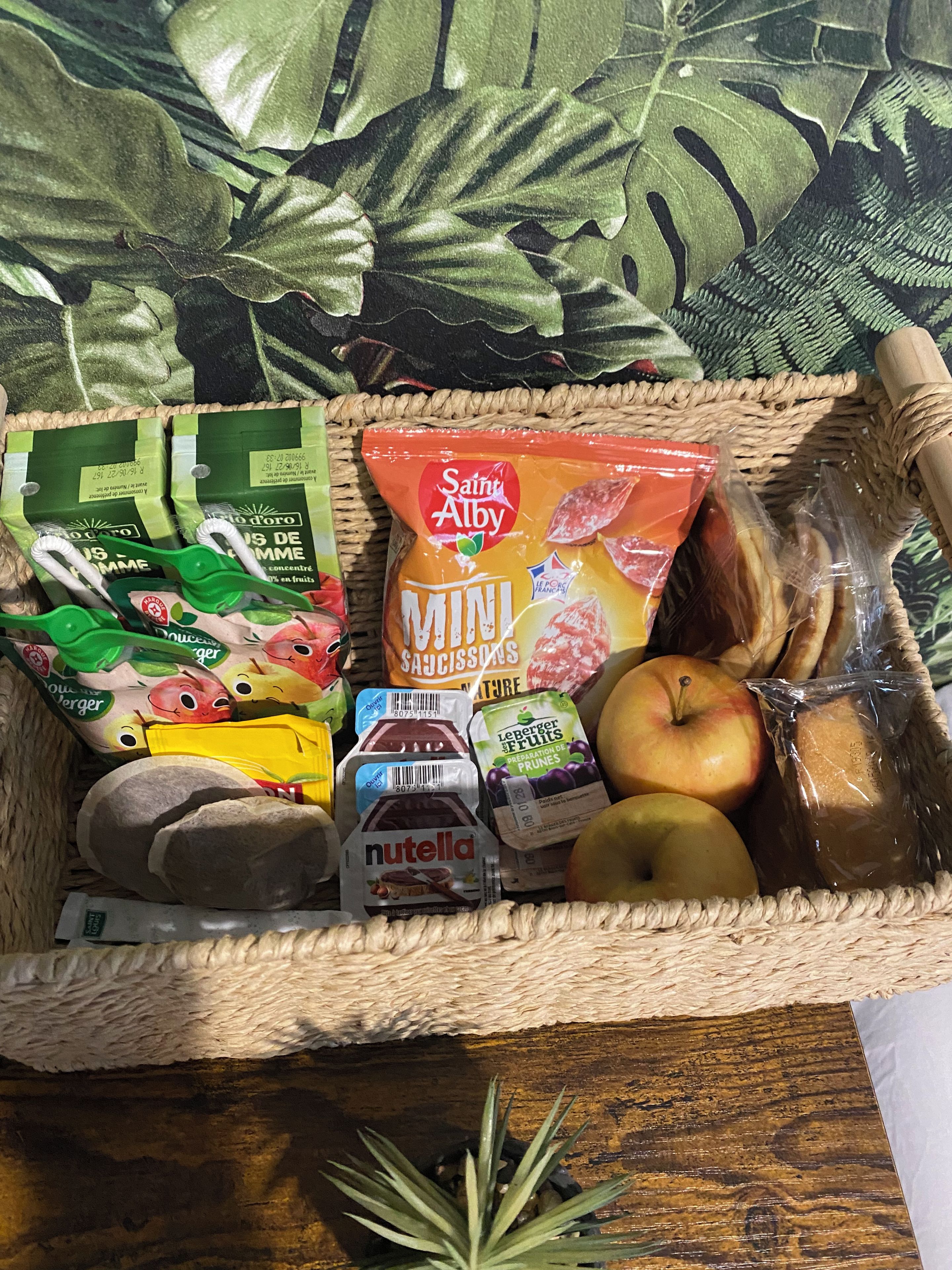 Een rieten mand gevuld met diverse etenswaren, waaronder een pakje Saint Alby Mini Saississons, een Nutella-reep en verschillende soorten fruit, zoals appels en bananen.