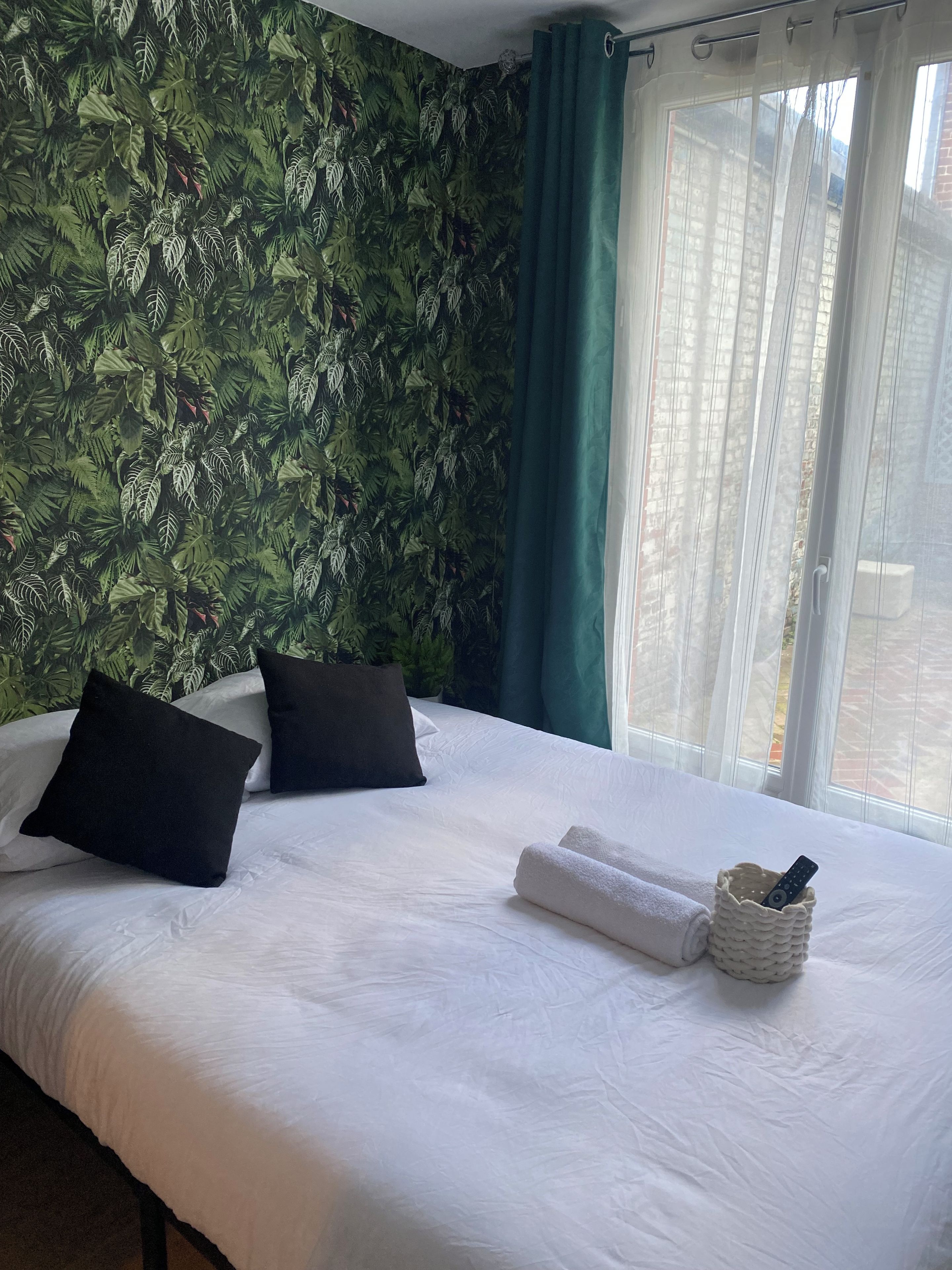 Een hotelkamer met een bed, kussens, een afstandsbediening in een mandje en uitzicht op gebouwen door een raam.