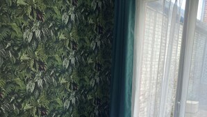 A hotel room with a bed, pillows, a remote control in a basket, and a view of buildings through a window.