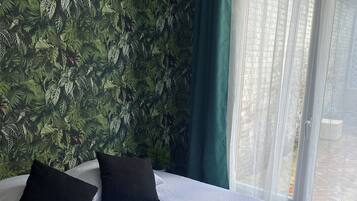Een hotelkamer met een bed, kussens, een afstandsbediening in een mandje en uitzicht op gebouwen door een raam.
