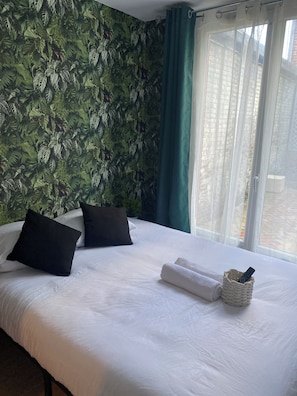 A hotel room with a bed, pillows, a remote control in a basket, and a view of buildings through a window.