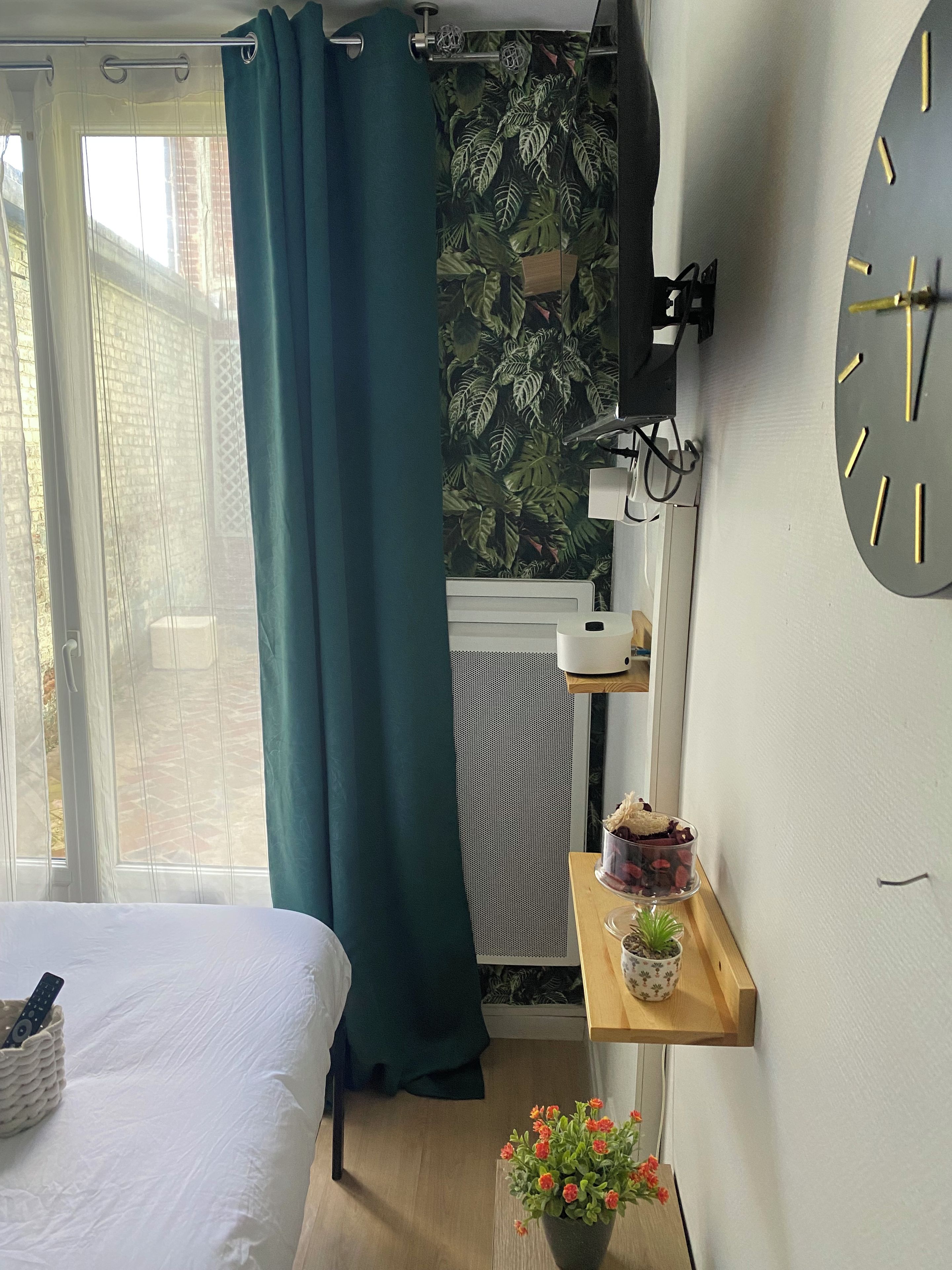 Een slaapkamer met een bed, een raam met gordijnen, een wandklok, een klein plankje met een plant en een radiator.