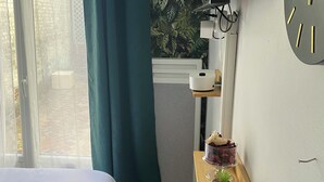 A bedroom with a bed, a window with curtains, a wall clock, a small shelf with a plant, and a radiator.
