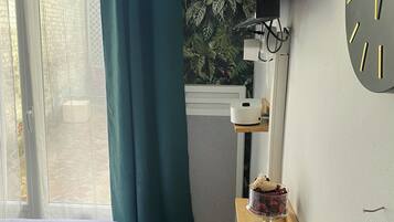 Een slaapkamer met een bed, een raam met gordijnen, een wandklok, een klein plankje met een plant en een radiator.