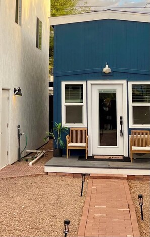 A building with a blue exterior, a white door, two windows, and two wooden benches outside. - Cozy, Modern Tiny Home, 1-bed, 1 bath - 10 minutes from UofA & other hotspots! (Tucson)
