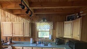 A wooden cabin kitchen with a stone tile floor, wooden cabinets, a sink, and a window with a view of greenery outside.