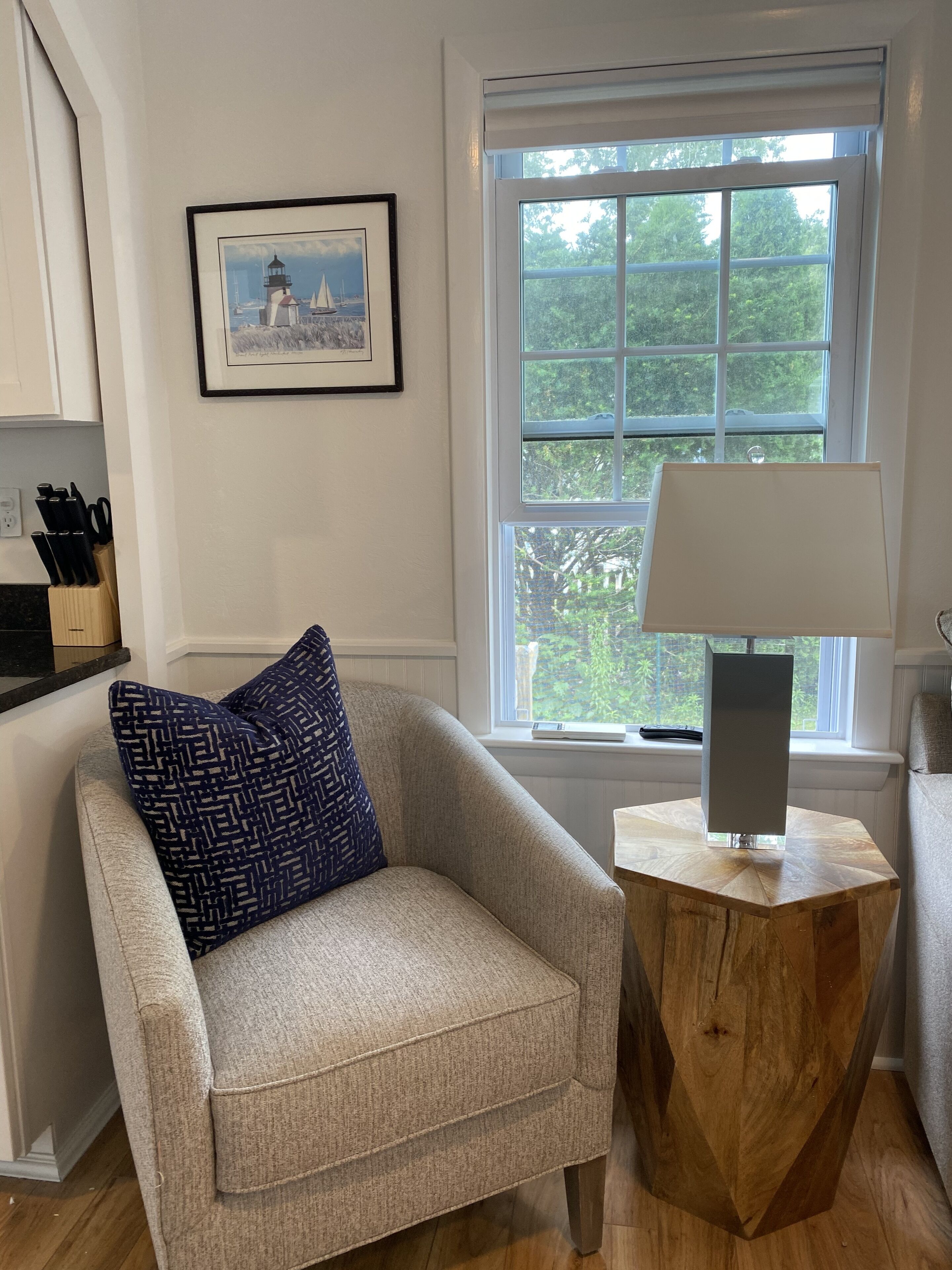 A cozy armchair with a patterned pillow, a small wooden side table with a lamp, a framed picture on the wall, and a window with a view of trees.