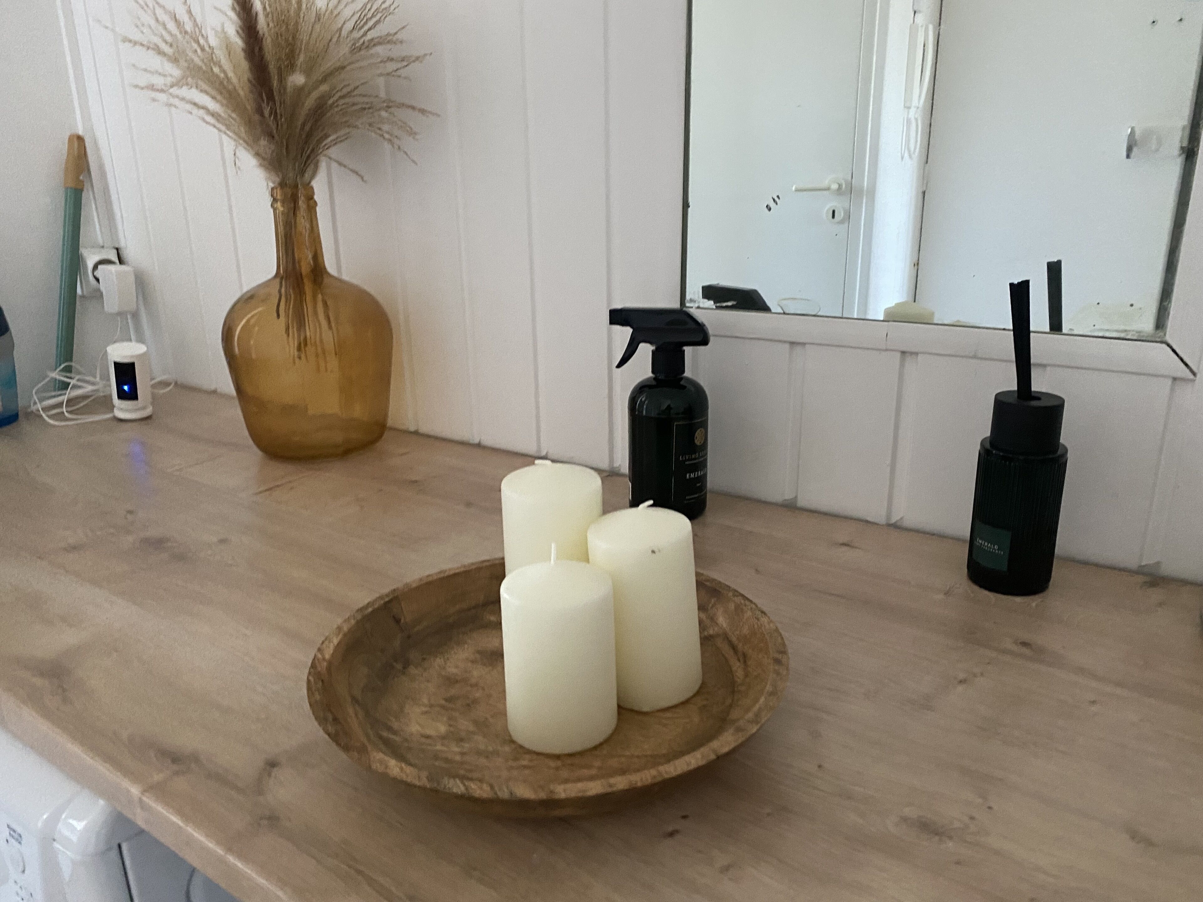 A wooden countertop with a round wooden tray holding three white candles, a spray bottle, and a vase with dried plants.