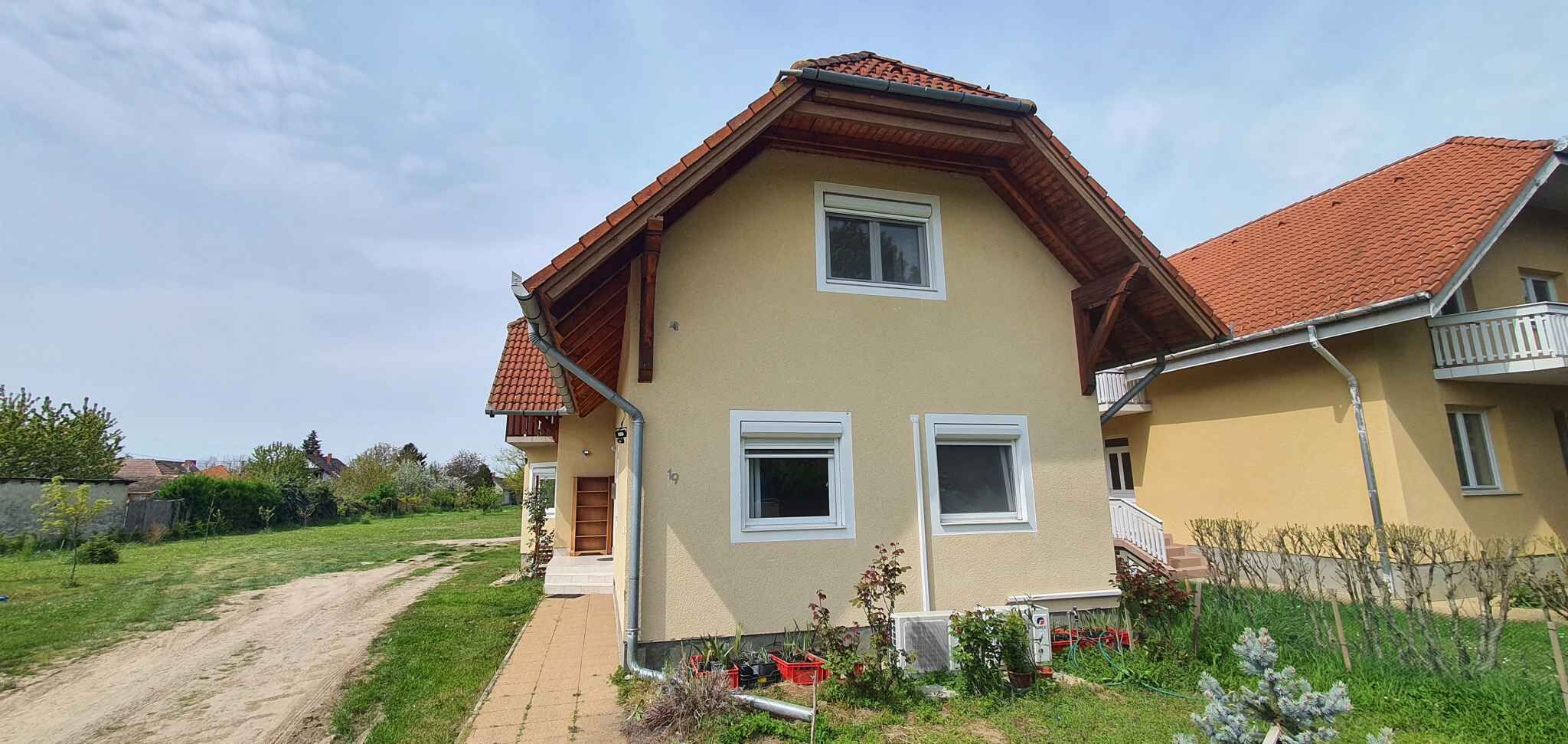 Uma casa de dois andares com telhado de telhas vermelhas, o número '19' visível na fachada e um caminho de paralelepípedos que leva à entrada.