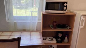 Een kleine keuken met een magnetron op een plank, een tafel met stoelen en een raam met uitzicht op groen.
