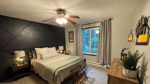 Un dormitorio con cama, mesitas de noche, escritorio, ventilador de techo, ventana con cortinas y una planta.