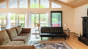 Ein geräumiges Wohnzimmer mit hohen Decken, großen Fenstern, einem schwarzen Ledersofa, einem beigen Sofa, einem gemusterten Teppich und einem Kamin aus Stein.