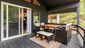Eine überdachte Terrasse mit Holztisch und -stühlen, eine Glastür zu einem Innenbereich und Blick ins Grüne.