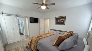 A bedroom with a bed, a ceiling fan, a TV, a door, and a rug. - Beautiful Modern Lake House Along Lake Oahe. (Pierre)