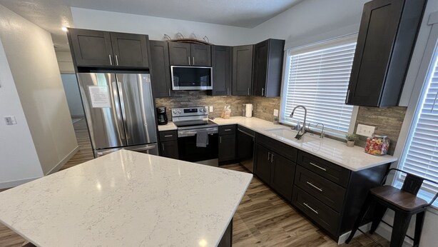 A modern kitchen with dark cabinets, a large island, stainless steel appliances, and a window with blinds. - Beautiful Modern Lake House Along Lake Oahe. (Pierre)