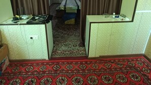 Une pièce avec une moquette à motifs, un lit recouvert d’une couvre-lit verte et blanche, une petite table avec une lampe et une fenêtre ornée d’un cadre décoratif.