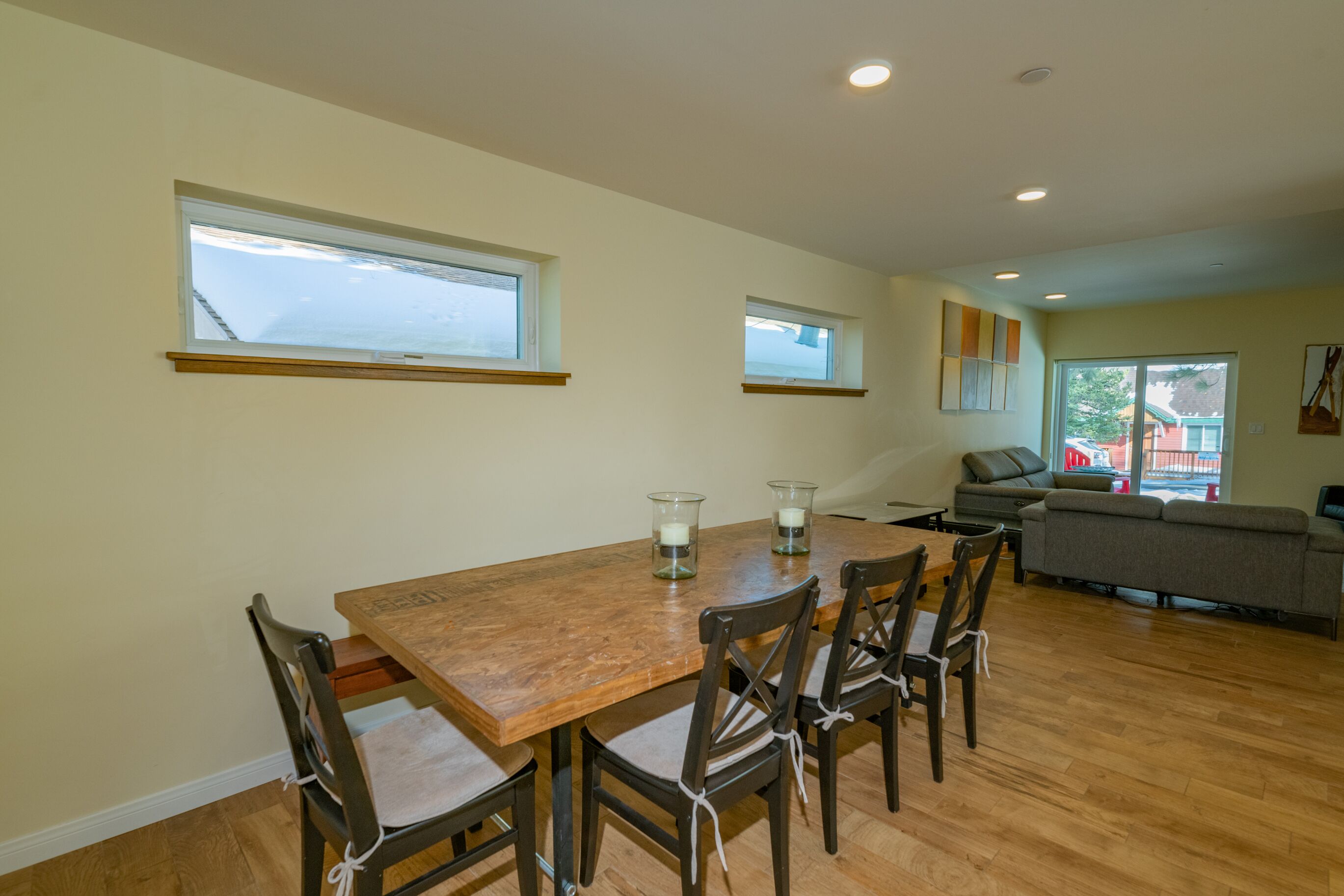 A dining area with a wooden table and chairs, two windows, a sofa, and a view of the outside.