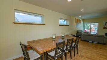 A dining area with a wooden table and chairs, two windows, a sofa, and a view of the outside.