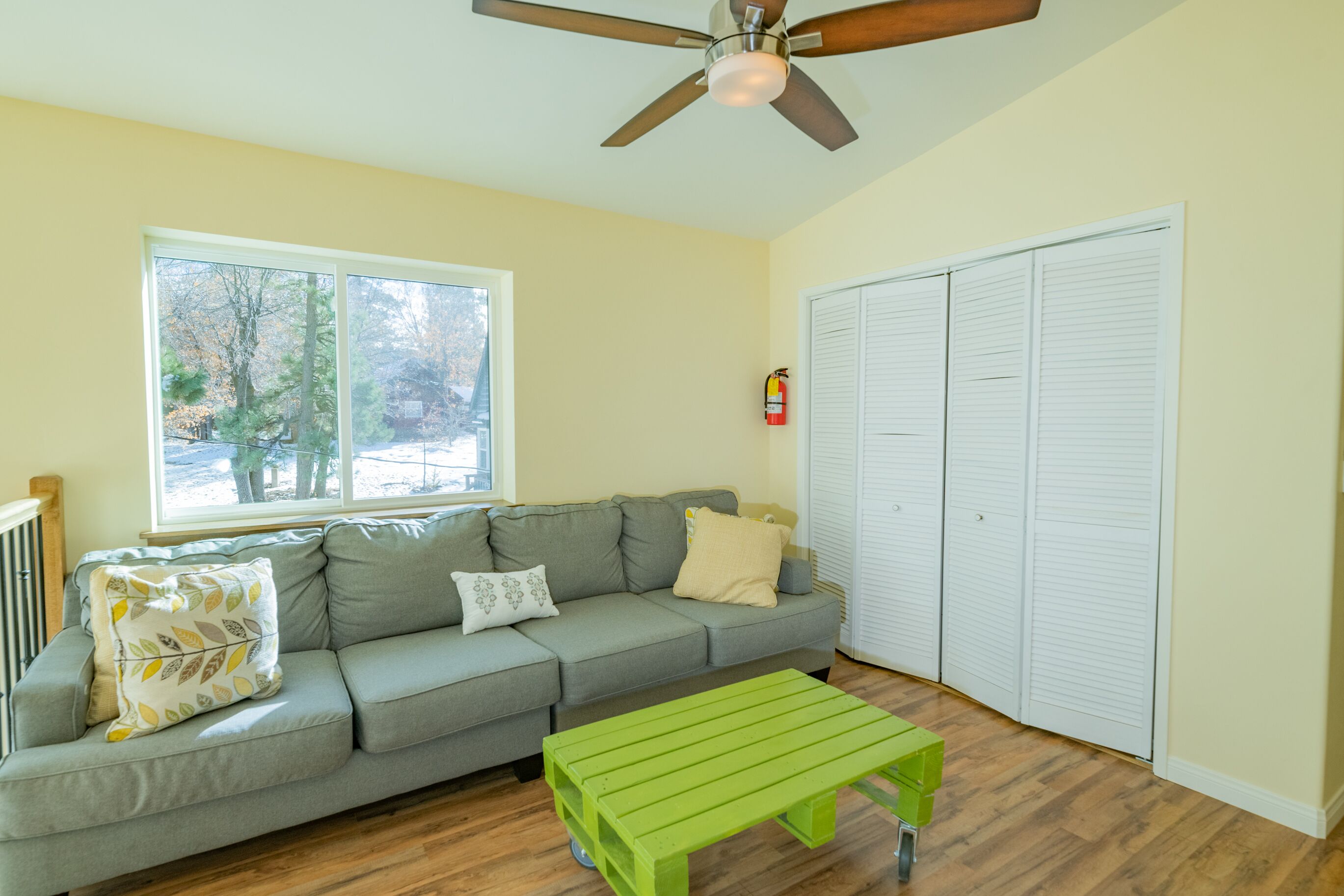 A living room with a gray sofa, a green coffee table, a ceiling fan, and a window with a view of trees.