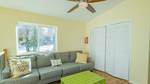 A living room with a gray sofa, a green coffee table, a ceiling fan, and a window with a view of trees. - Modern Times (Big Bear Lake)