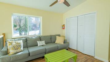 A living room with a gray sofa, a green coffee table, a ceiling fan, and a window with a view of trees.