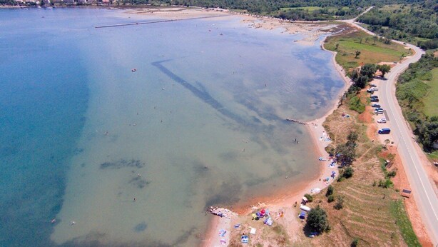 An aerial view of a coastal area with a clear beach, a road along the coast, and a distant town.