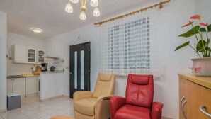 A modern living room with a beige and red leather sofa, a wooden coffee table with fruit, a kitchen area with white cabinets, and a window with lace curtains.