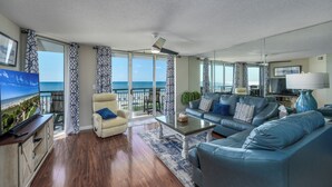 A spacious living room with a large flat-screen TV, leather sofas, a coffee table, and a view of the ocean through large windows. - Windy Hill Dunes-Oceanfront 3BD-Family Friendly (North Myrtle Beach)