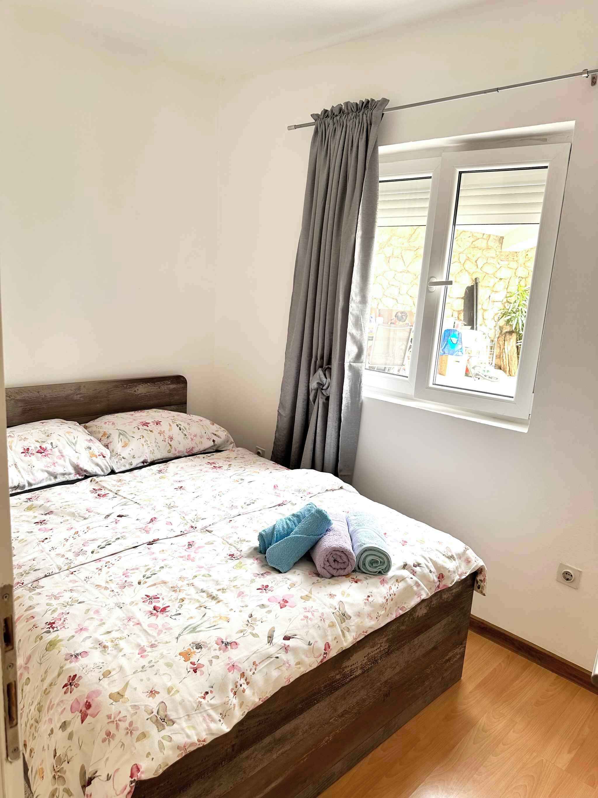 Een slaapkamer met een bed, bloemenbedrukking, een raam met gordijnen en uitzicht op buiten.