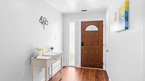 A hallway with a wooden door, a white wall with a heart-shaped decoration, a small table with a vase, and a painting on the wall. - NEW IMMACULATE and quiet 4br Retreat between SAV DownTown & Tybee Island (Savannah)