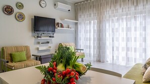 Una sala de estar con televisor de pantalla plana, dos sofás, una mesa de centro con un centro de mesa de verduras y un amplio ventanal con cortinas translúcidas.