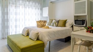 Un dormitorio con cama, escritorio, silla, microondas y un gran ventanal con cortinas translúcidas.