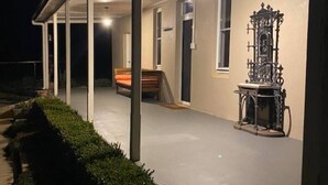 A covered outdoor area with a bench, a decorative metal piece, and a pathway lined with bushes. - Authentic Farm Cottage (Cowra)
