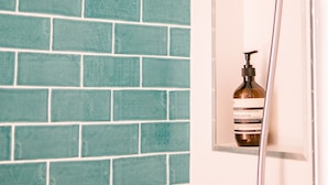 A bathroom with a teal brick wall, a showerhead, and a bottle of shampoo in a mirror. - Ocean Views at Sandrift on Nobby Beach (Gold Coast)
