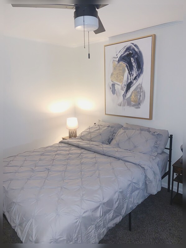 A bedroom with a bed, a ceiling fan, a wall-mounted abstract painting, and a bedside table with a lamp. - Two BR - Convenient Oasis (Deer Park)