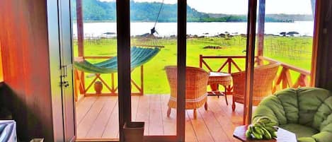Una habitación con piso de madera, una hamaca, sillas de mimbre y vistas a un lago y campos verdes.