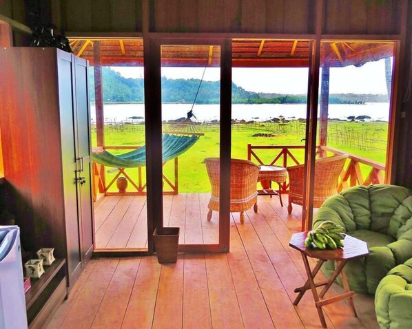 A room with a wooden floor, a hammock, wicker chairs, and a view of a lake and green fields. - Simeulue Surf Camp (Sinabang)