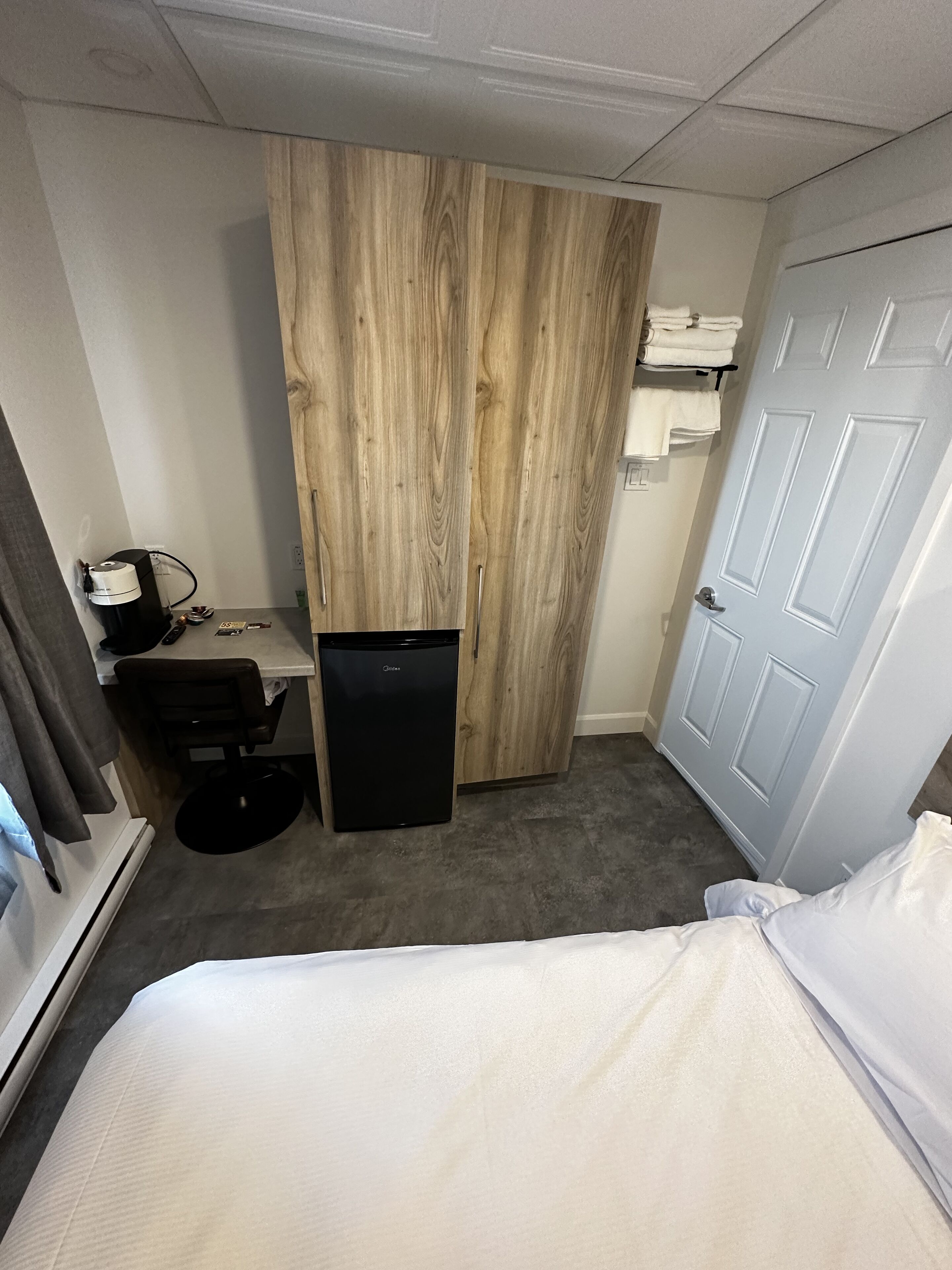 A hotel room with a bed, a desk with a computer, a small refrigerator, a wardrobe, and a door.