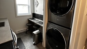 Eine kompakte Küche mit integriertem Waschtrockner, einem kleinen Barhocker und einem Fenster mit Jalousien.