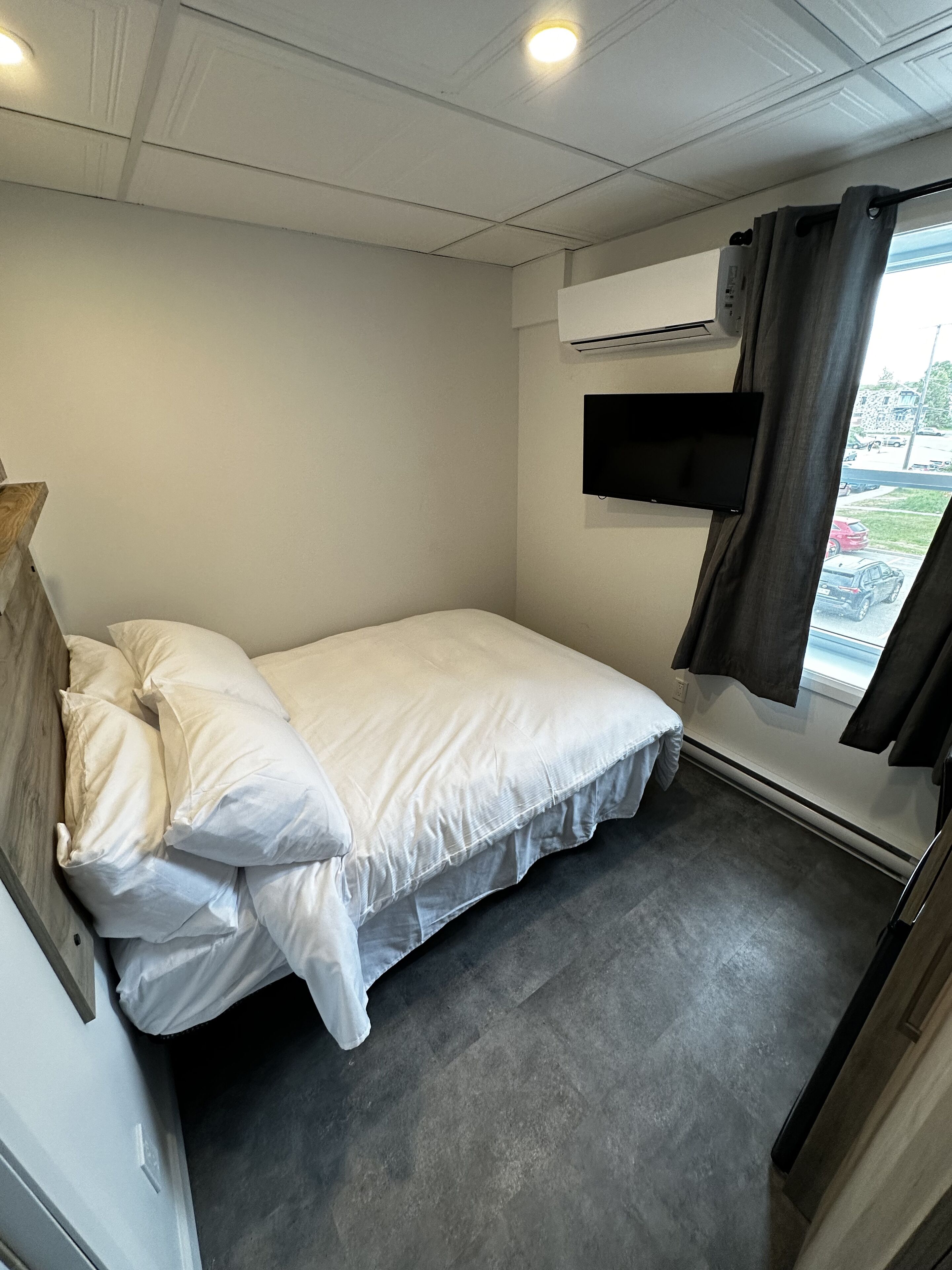 A small, single-bed room with a grey carpet, white walls, a mounted TV, and a window with curtains.