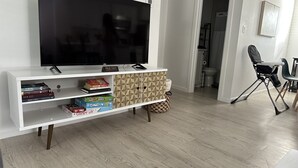 Een woonkamer met een flatscreen televisie, een boekenkast met boeken en gezelschapsspellen, een kleine tafel met een stoel, en een kinderstoel.
