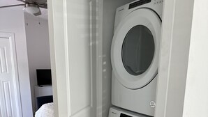 Een wasserette met twee witte voorladers, een plafondventilator en een deur naar een slaapkamer met een bed en een televisie.